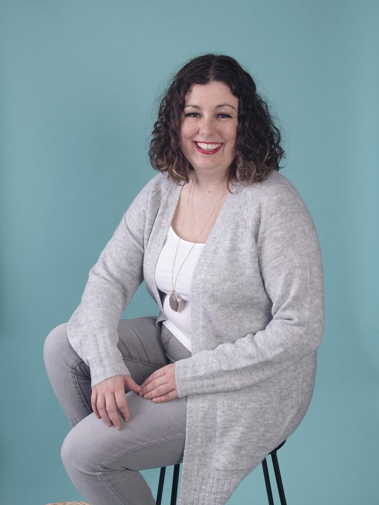 Female life coach dressed in grey sits on a stool in front of a blue backdrop and smiles. 