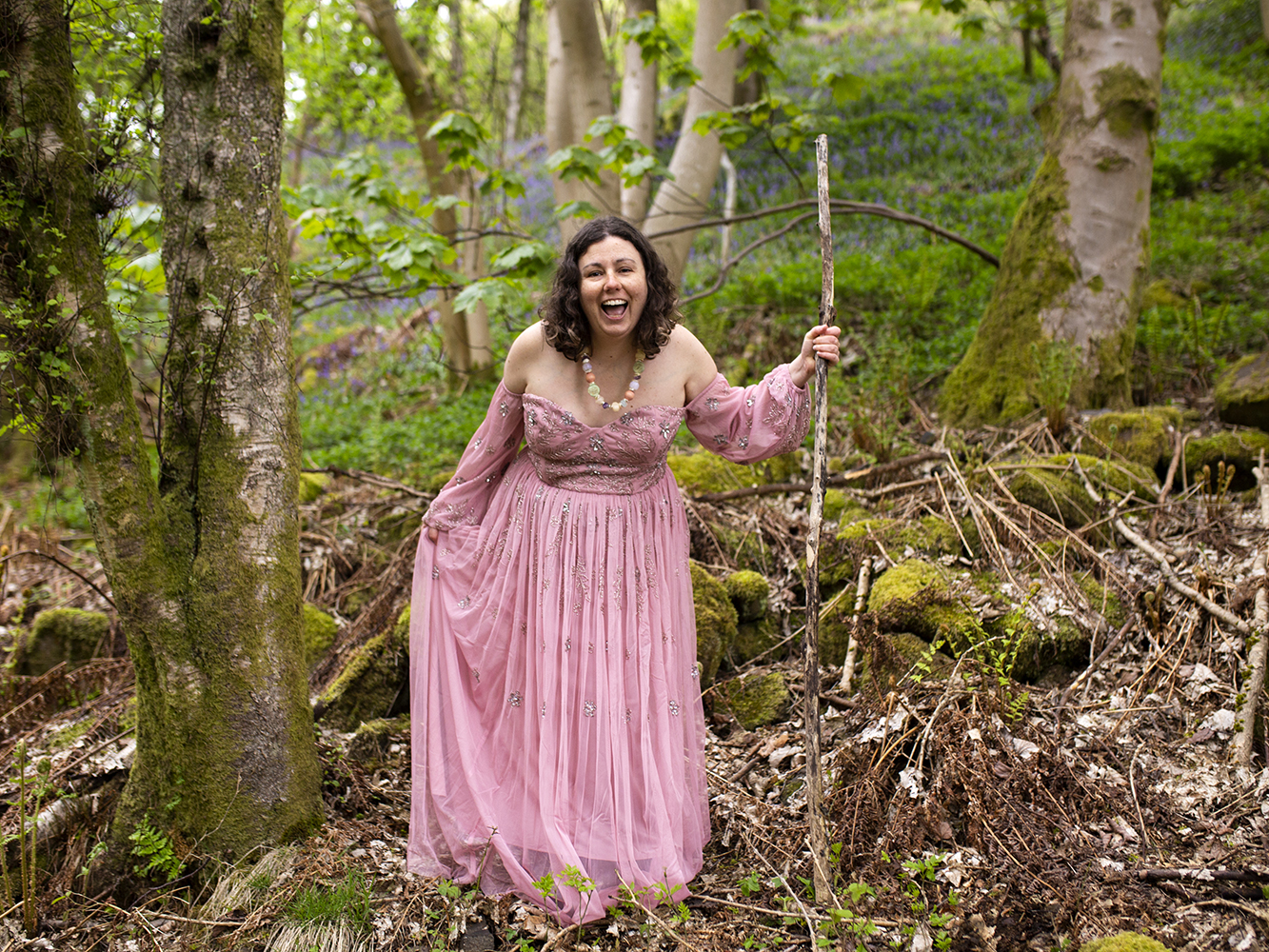 Woman in a pink gown holds a wooden staff and stands in the forest. Her mouth is open and smiling