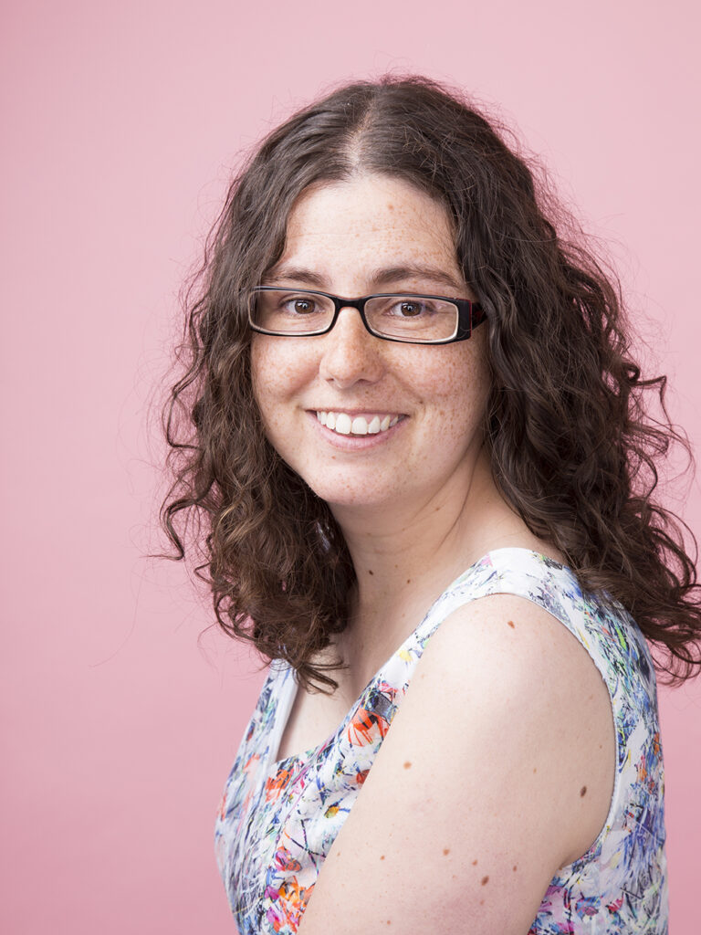 Headshot of a smiling woman wearing glasses and a flowery dress on a pink background